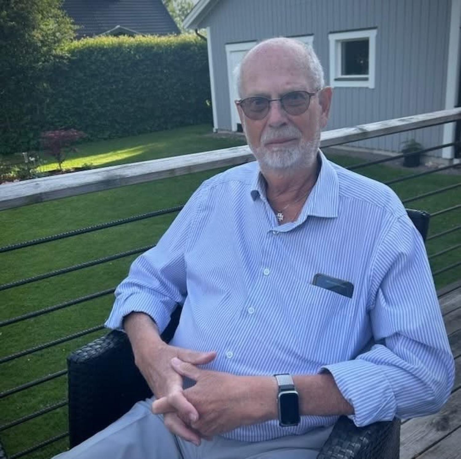 En äldre man med skägg och glasögon sitter på en utestol utomhus, klädd i en randig skjorta och med en klocka på armen, med en gräsmatta och ett grått skjul i bakgrunden.