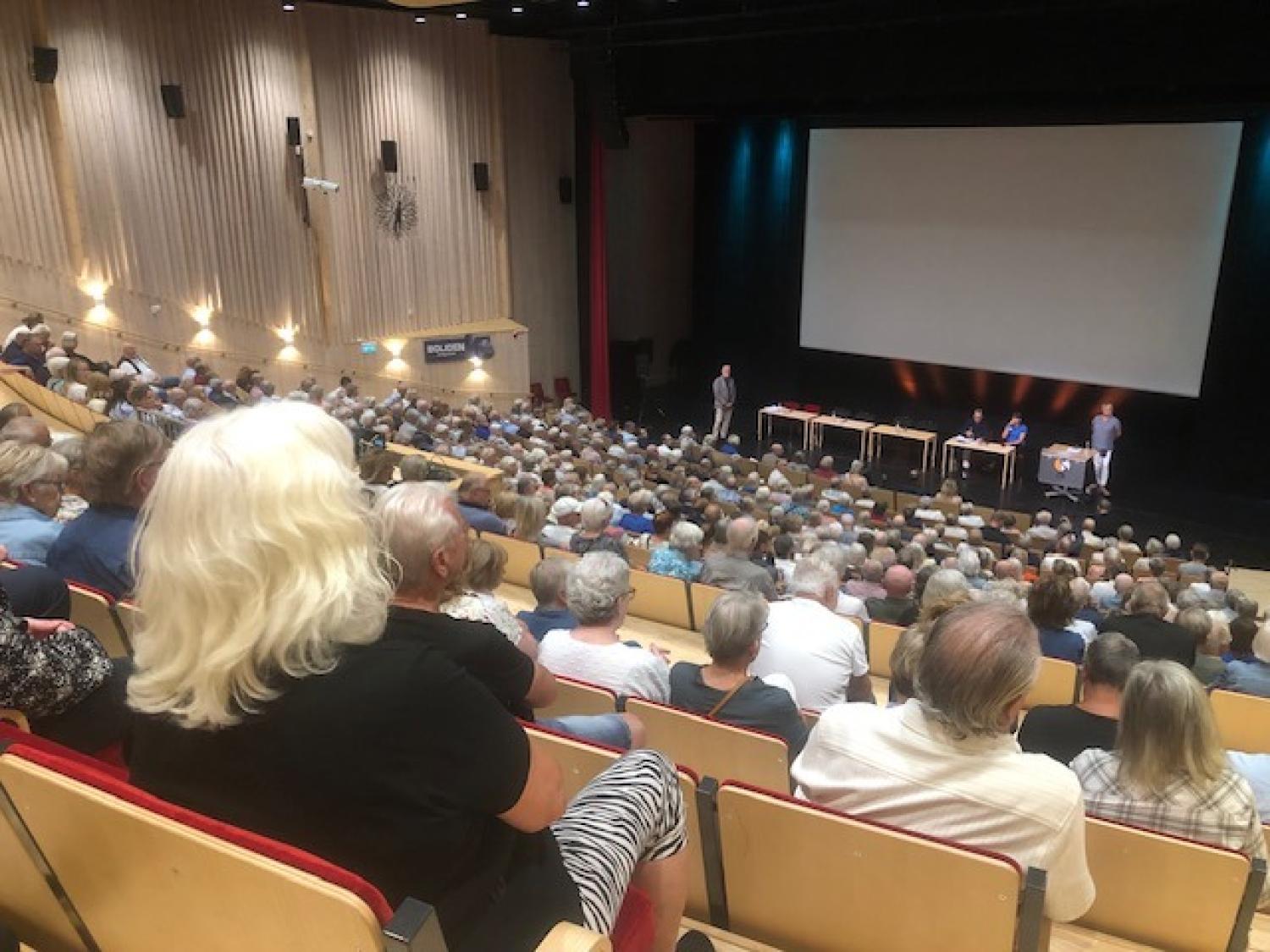 En stor publik sitter i en sal med teateruppställning vänd mot en scen där fyra personer sitter vid bord och en person står bredvid en skärm.