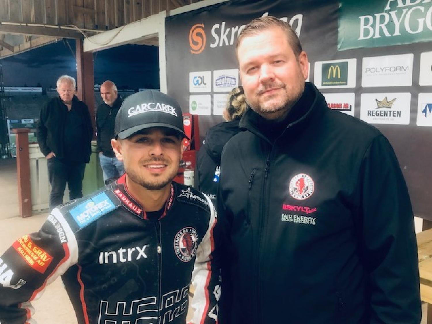 Två män står bredvid varandra inomhus; den ene har på sig en racerdräkt och en keps, den andre har på sig en svart jacka. Sponsorslogotyper syns på deras kläder och på väggen bakom dem.