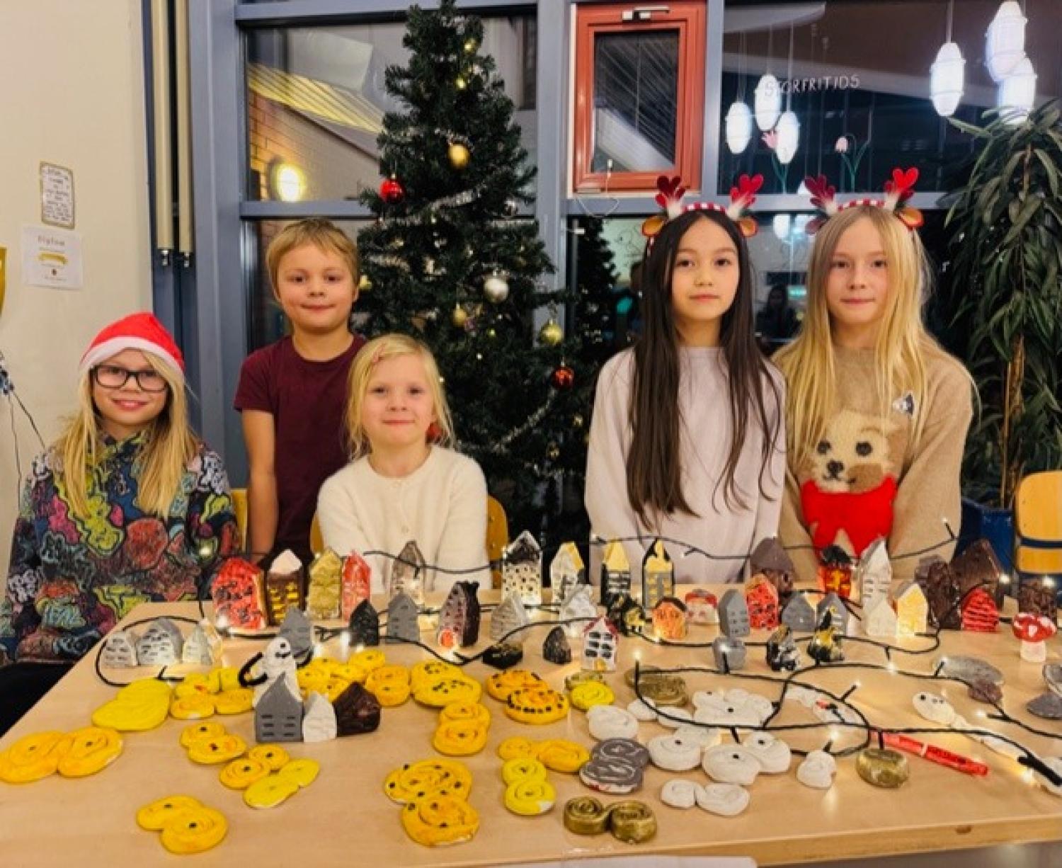 Fem barn står bakom ett bord med handgjorda julpyssel och dekorationer, med en dekorerad julgran i bakgrunden.