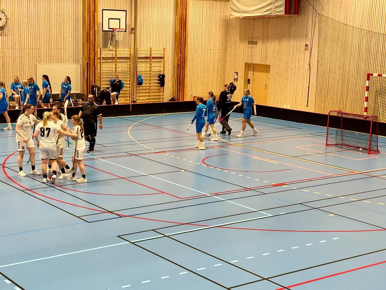Två damlag i innebandy, ett i vitt och ett i blått, spelar en match i en inomhushall med ett rött mål och träväggar.