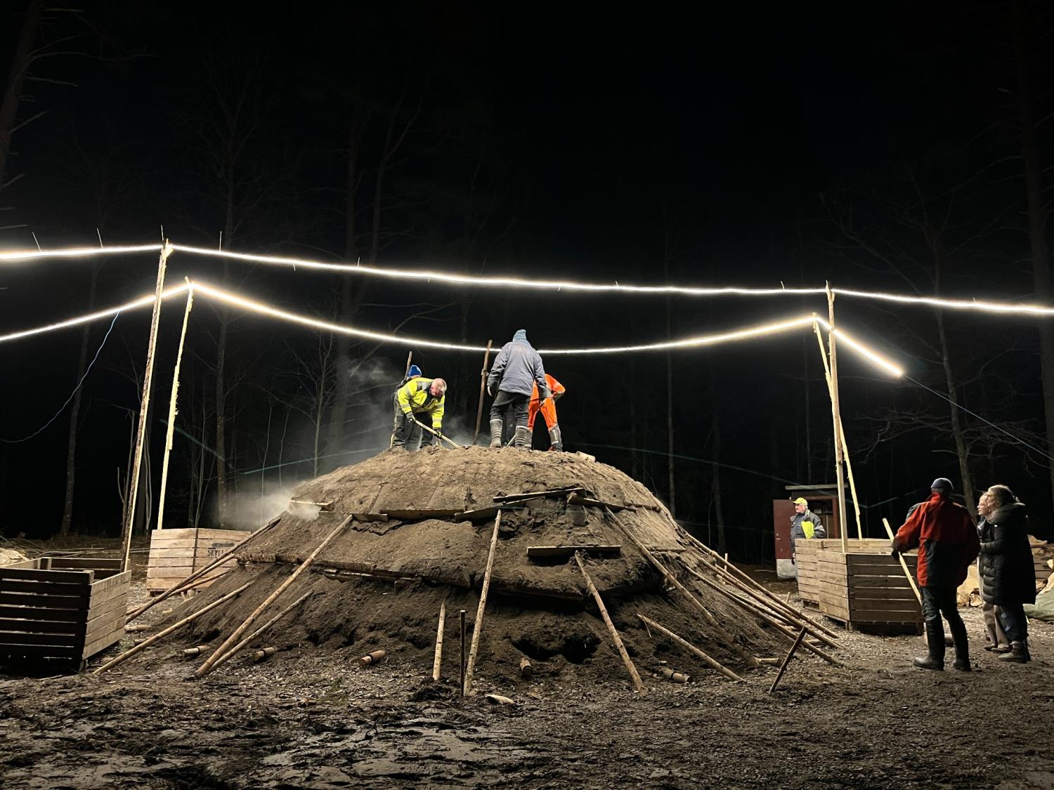 Flera personer arbetar på och runt en stor, kupolformad jordhög under starka lampor på natten, med träplankor och stolpar som stöd för strukturen.