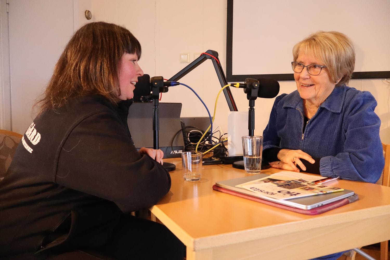Två kvinnor sitter mitt emot varandra vid ett bord med mikrofoner, inspelningsutrustning, vattenglas och en tidning, vilket tyder på en podcast eller en intervju.