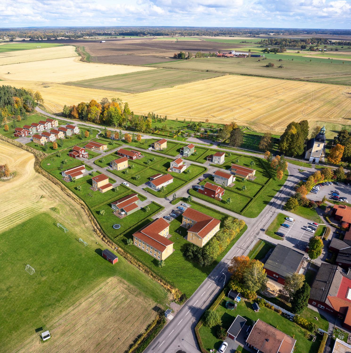 Flygfoto över ett litet bostadsområde med hus med röda tak, omgivet av fält och vägar på landsbygden.