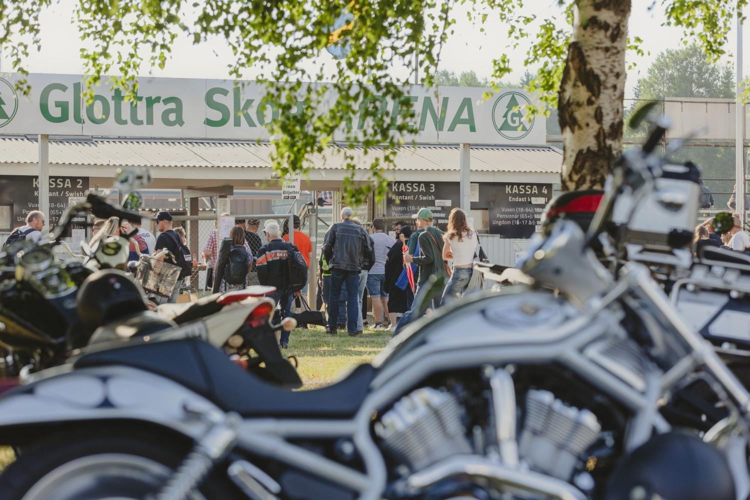 Människor samlas utanför Glötta Skog Arena vid biljettkioskerna, med flera parkerade motorcyklar i förgrunden och träd som delvis skymmer sikten.