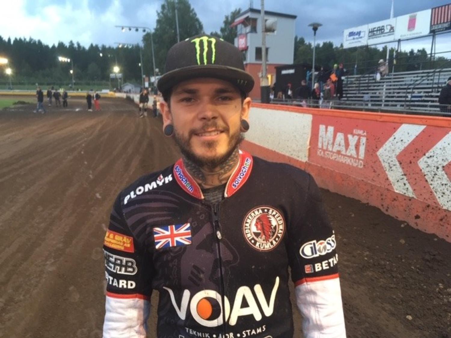 En man som bär en patch med den brittiska flaggan och en racerdräkt med sponsorlogotyper står på en grusbana, med tomma läktare och träd i bakgrunden.