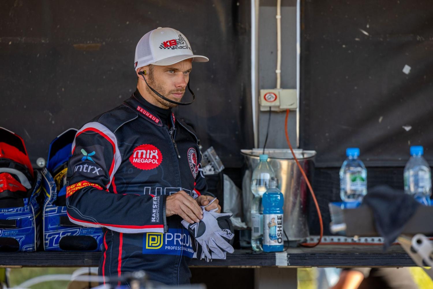 En racerbilsförare i racingdräkt och keps står inomhus med handskar i handen och vattenflaskor och racingutrustning i bakgrunden.