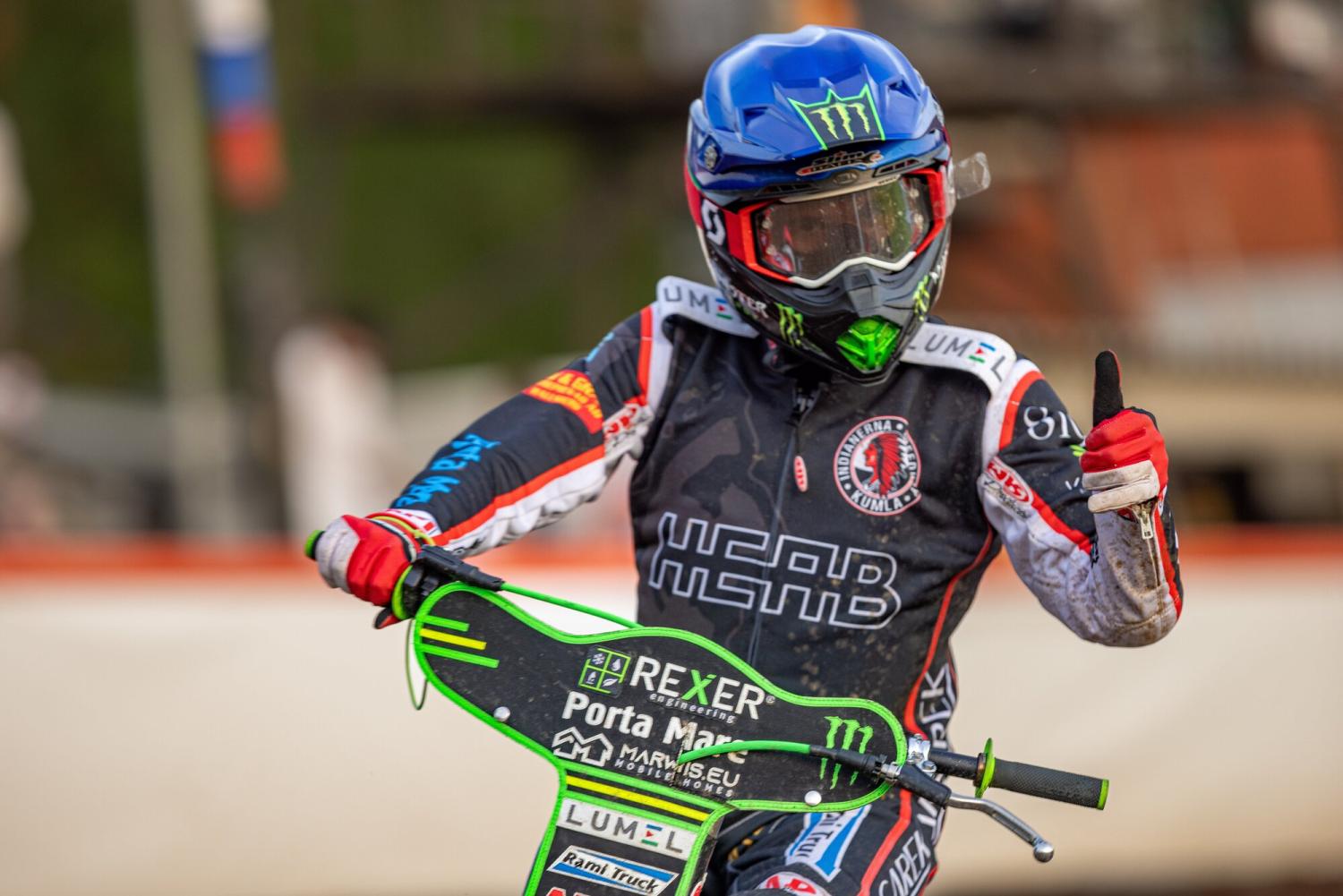 En motocrossförare i full utrustning och hjälm ger tummen upp när han sitter på en grön och svart cykel under ett lopp eller evenemang.