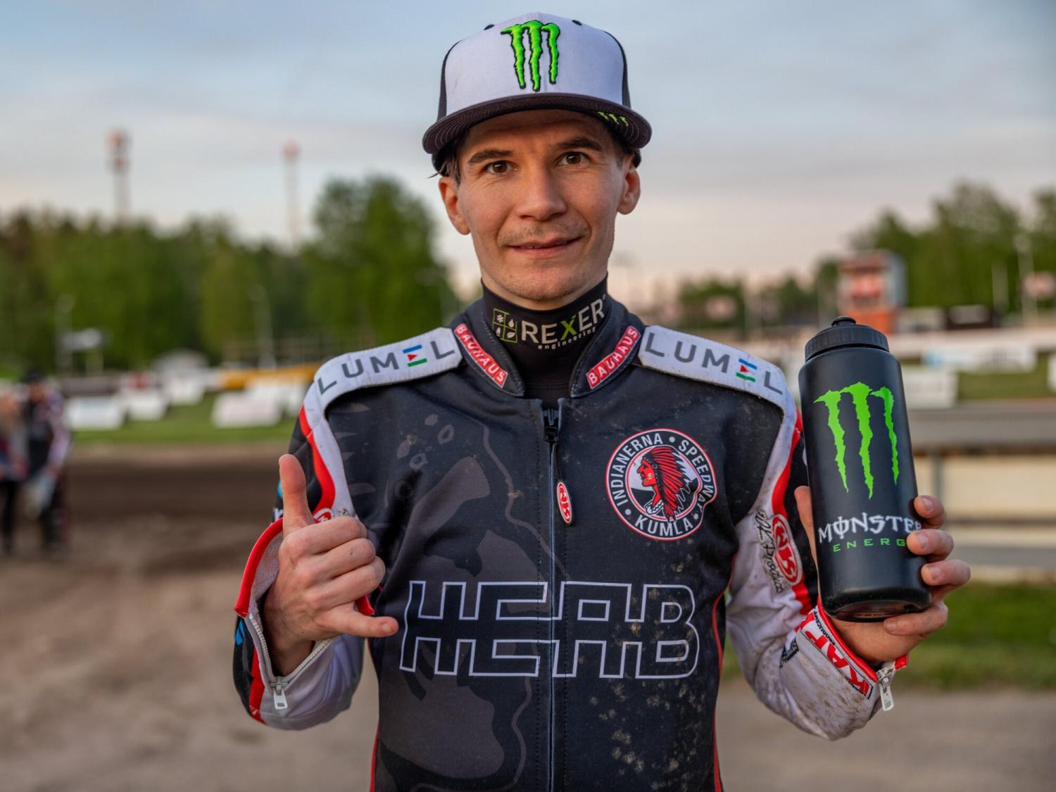 En person i racingutrustning håller en Monster Energy-drinkflaska. De står på en racerbana med träd i bakgrunden och gör en gest med händerna.