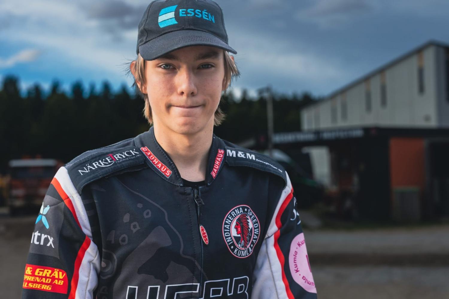 En ung person i racingdräkt och keps står utomhus framför en byggnad och tittar in i kameran med ett neutralt uttryck.