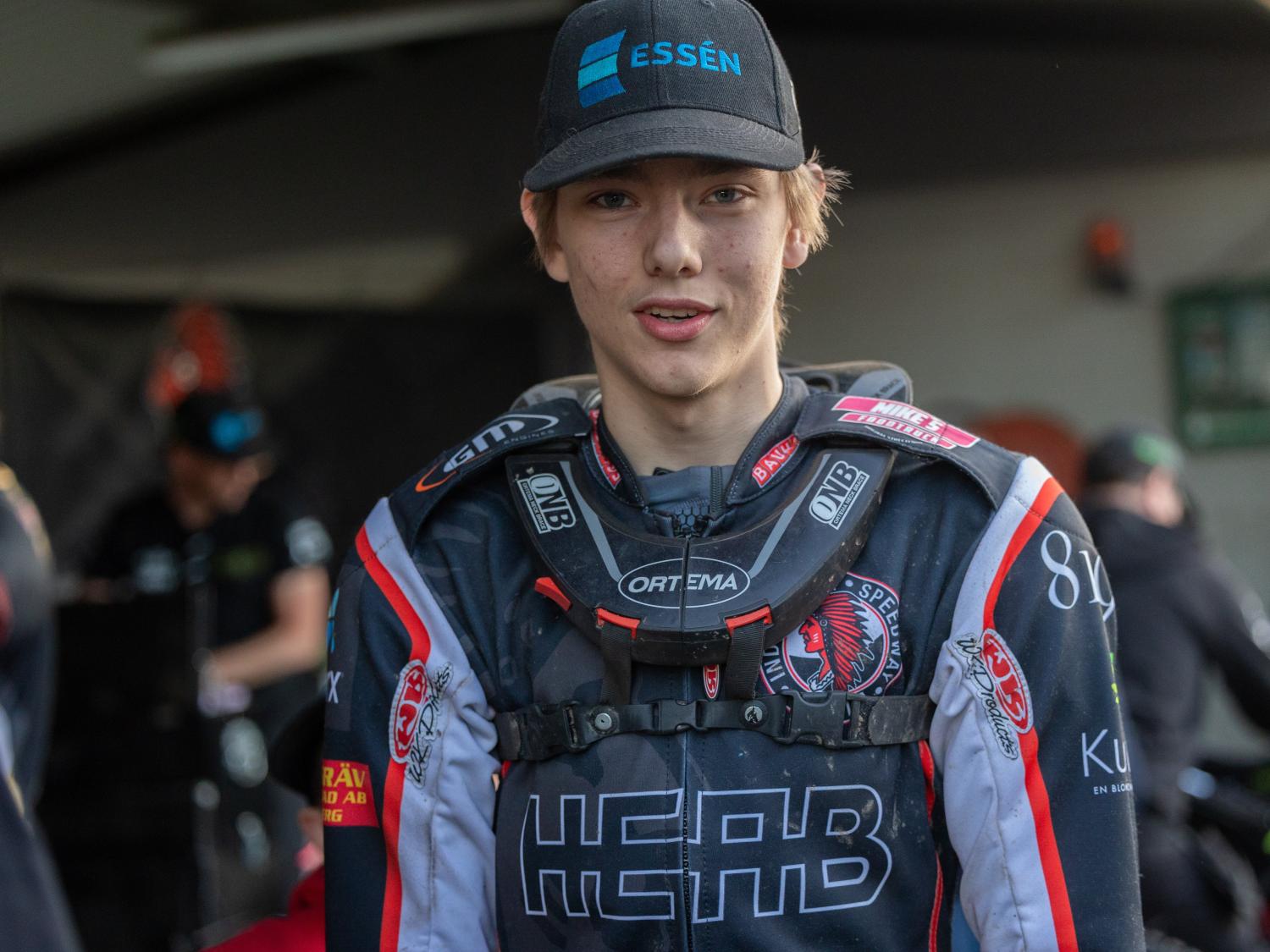 En ung manlig speedwayförare iförd tävlingskläder, svart keps och skyddsutrustning står inomhus och tittar in i kameran.