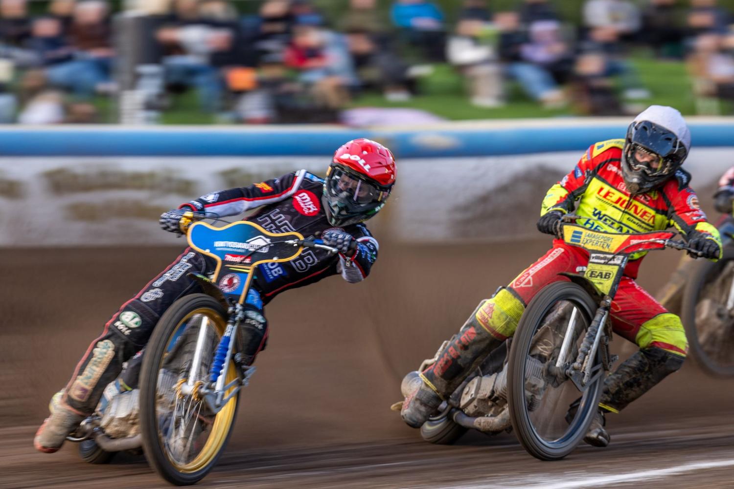Två speedwayförare i färgglad utrustning lutar sig in i en kurva på en grusbana, med suddiga åskådare i bakgrunden.