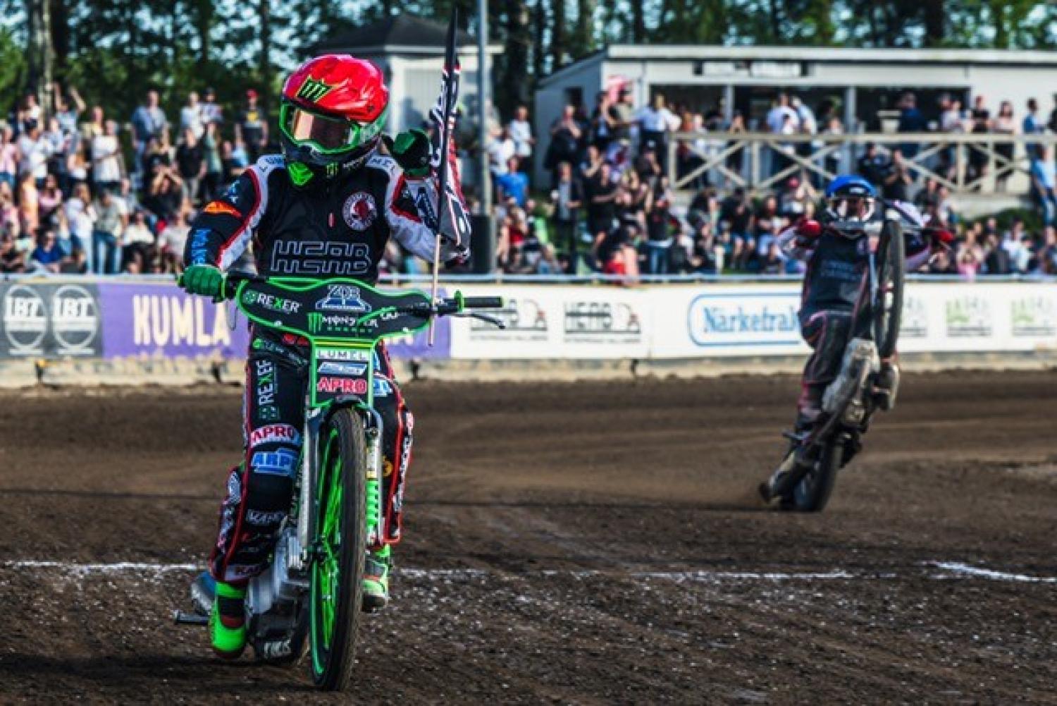 Två speedwaymotorcyklister på en grusbana; den ena kör upprätt och håller i en liten flagga, medan den andra utför en wheelie i bakgrunden med åskådare som tittar på.