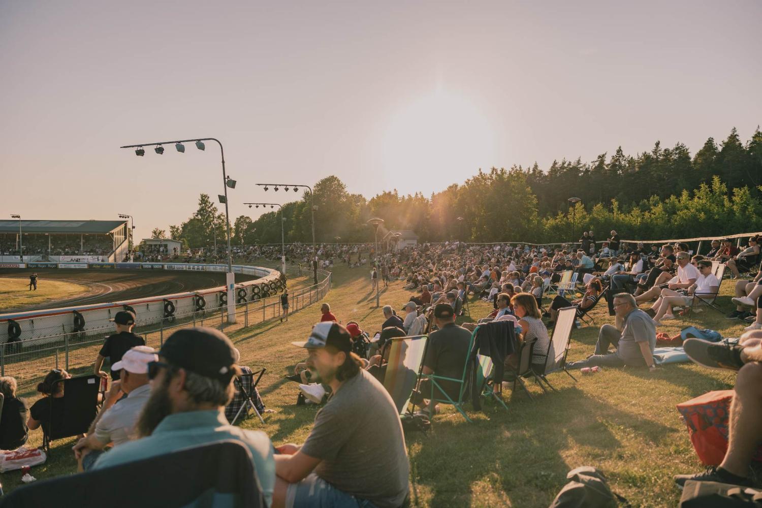 En stor folkmassa sitter på en gräsbevuxen sluttning och tittar på ett lopp på en utomhusbana under solnedgången, med träd och stadionbelysning i bakgrunden.