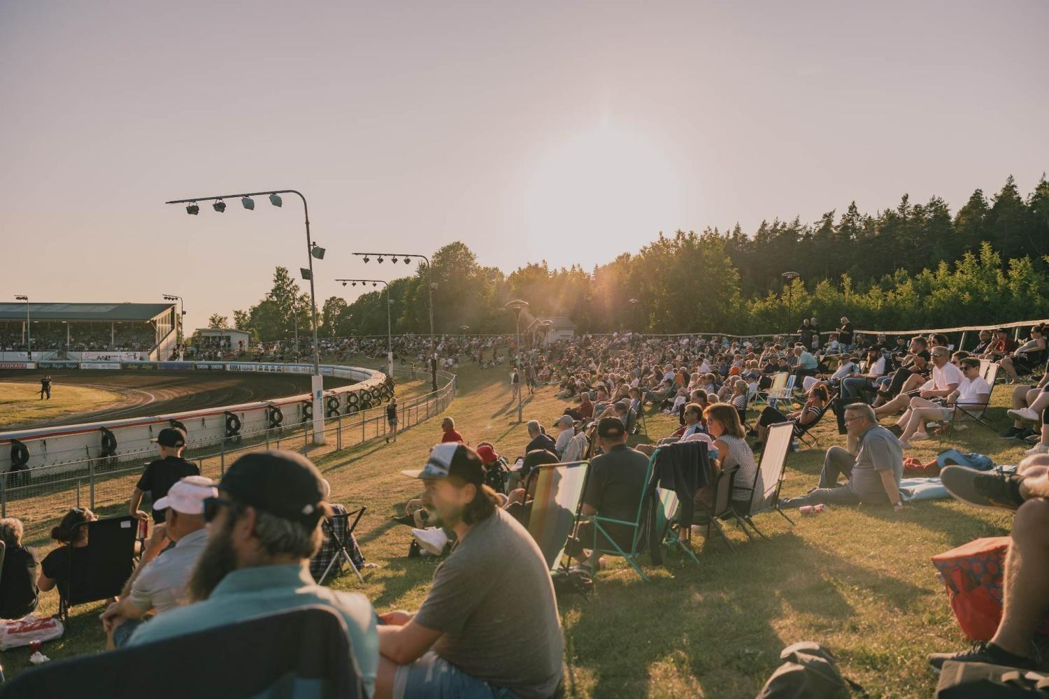 En stor publik sitter på en grässlänt och tittar på ett lopp på en speedwaybana i solnedgången, med träd och stadionljus i bakgrunden.