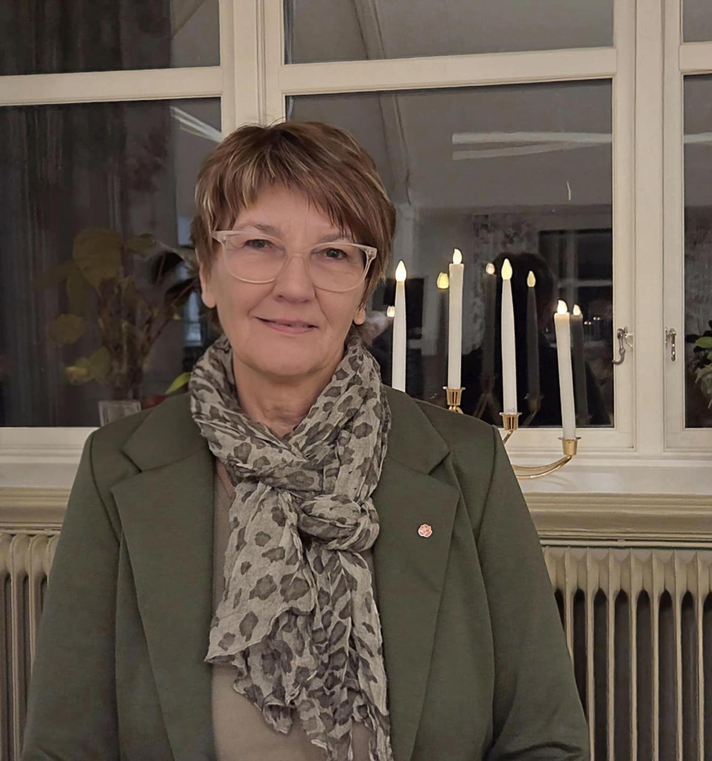 En kvinna med kort hår och glasögon, klädd i grön kavaj och mönstrad scarf, står inomhus framför ett fönster med en tänd ljusstake i bakgrunden.