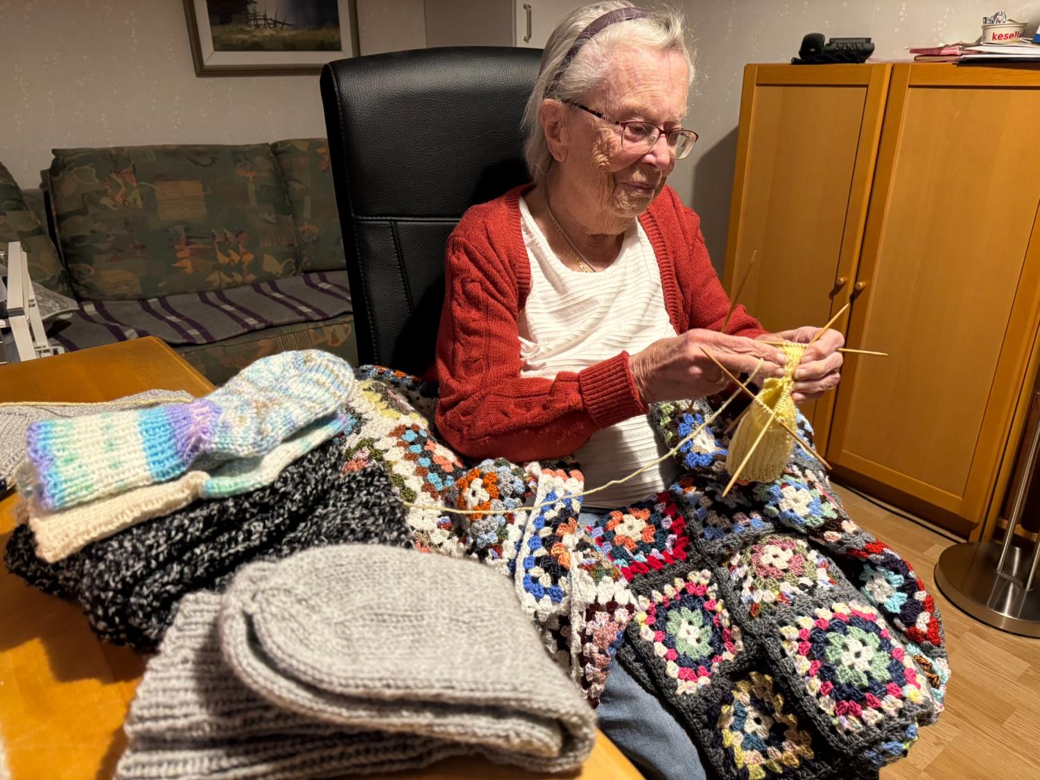 En äldre kvinna med glasögon sitter i en stol och stickar, omgiven av virkade filtar och vikta stickade bitar på ett bord i ett mysigt rum.