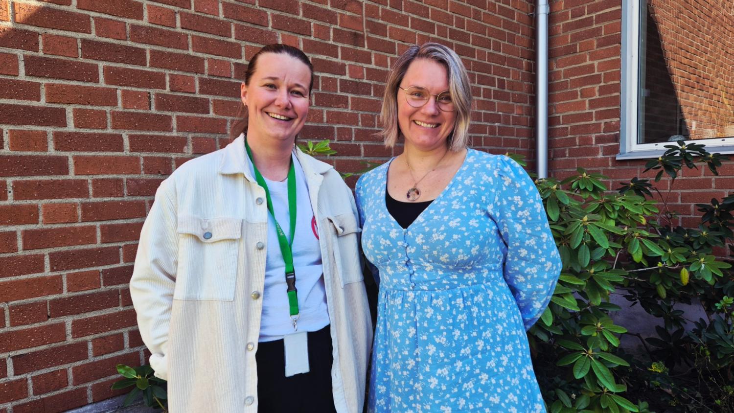 Två kvinnor står och ler framför en tegelvägg, den ena bär en vit jacka och nyckelband, den andra en blåblommig klänning.