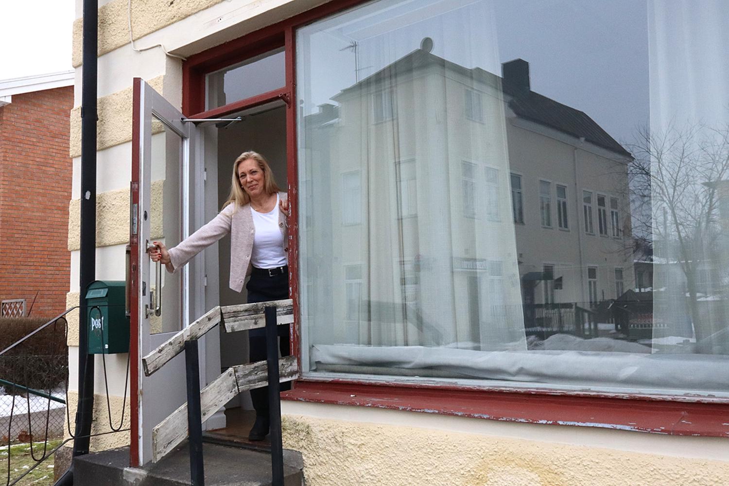 En kvinna står i en öppen dörröppning på en liten veranda utanför en byggnad med stora fönster, håller dörren öppen och tittar framåt.