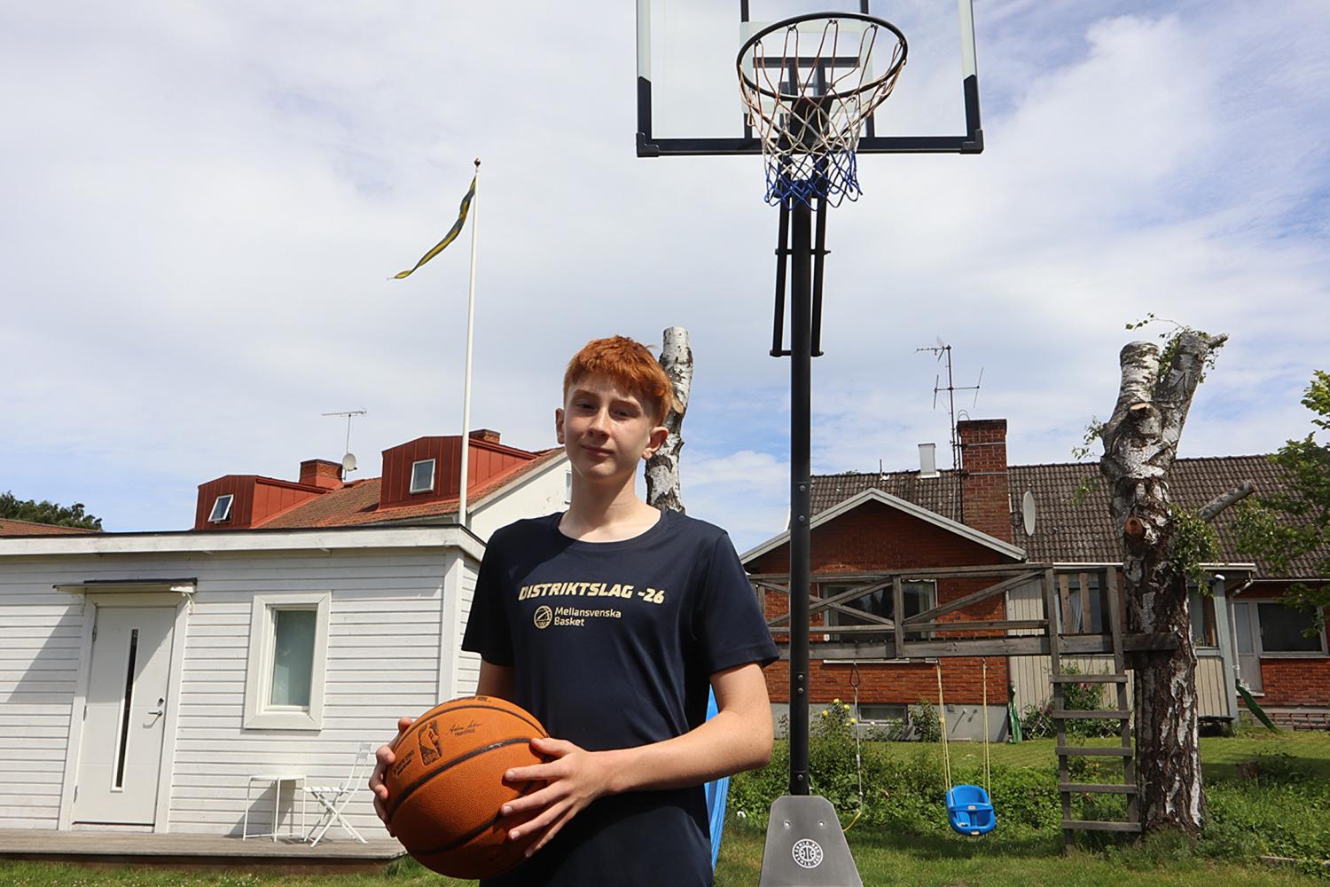 En tonårspojke som håller en basketboll står framför en utomhusbasketkorg i en trädgård, med ett vitt skjul och hus av rött tegel i bakgrunden.