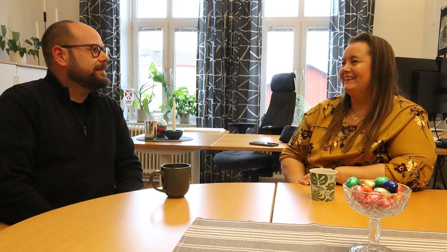 Två personer sitter vid ett runt bord på ett kontor och ler och pratar, med muggar och en skål med godis på bordet.