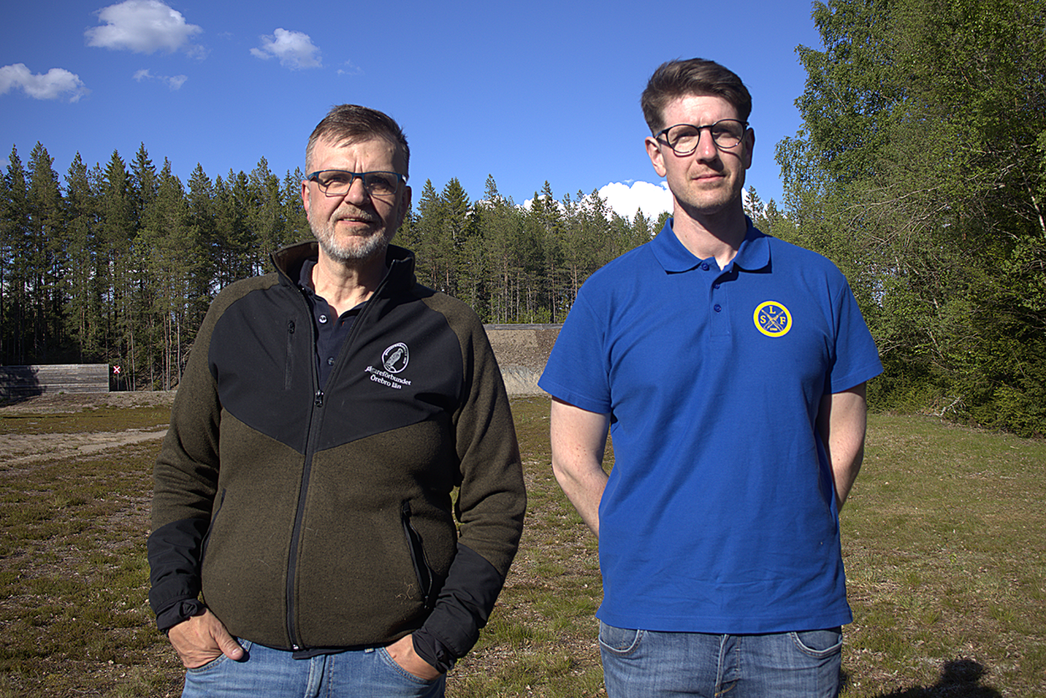 Två män står utomhus på en gräsmatta med träd och en blå himmel i bakgrunden; den ene bär en mörk jacka, den andre en blå polotröja med en cirkelformad logotyp.