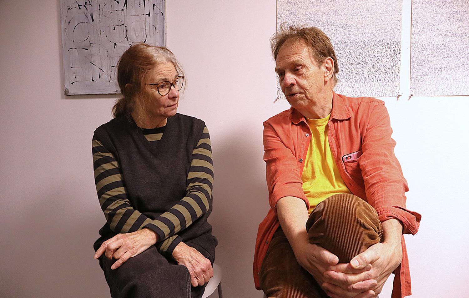 Två äldre vuxna sitter inomhus, vända mot varandra och samtalar. Kvinnan bär glasögon, randig skjorta och mörk väst, mannen har en orange skjorta över en gul T-shirt. På väggen hänger en konst.