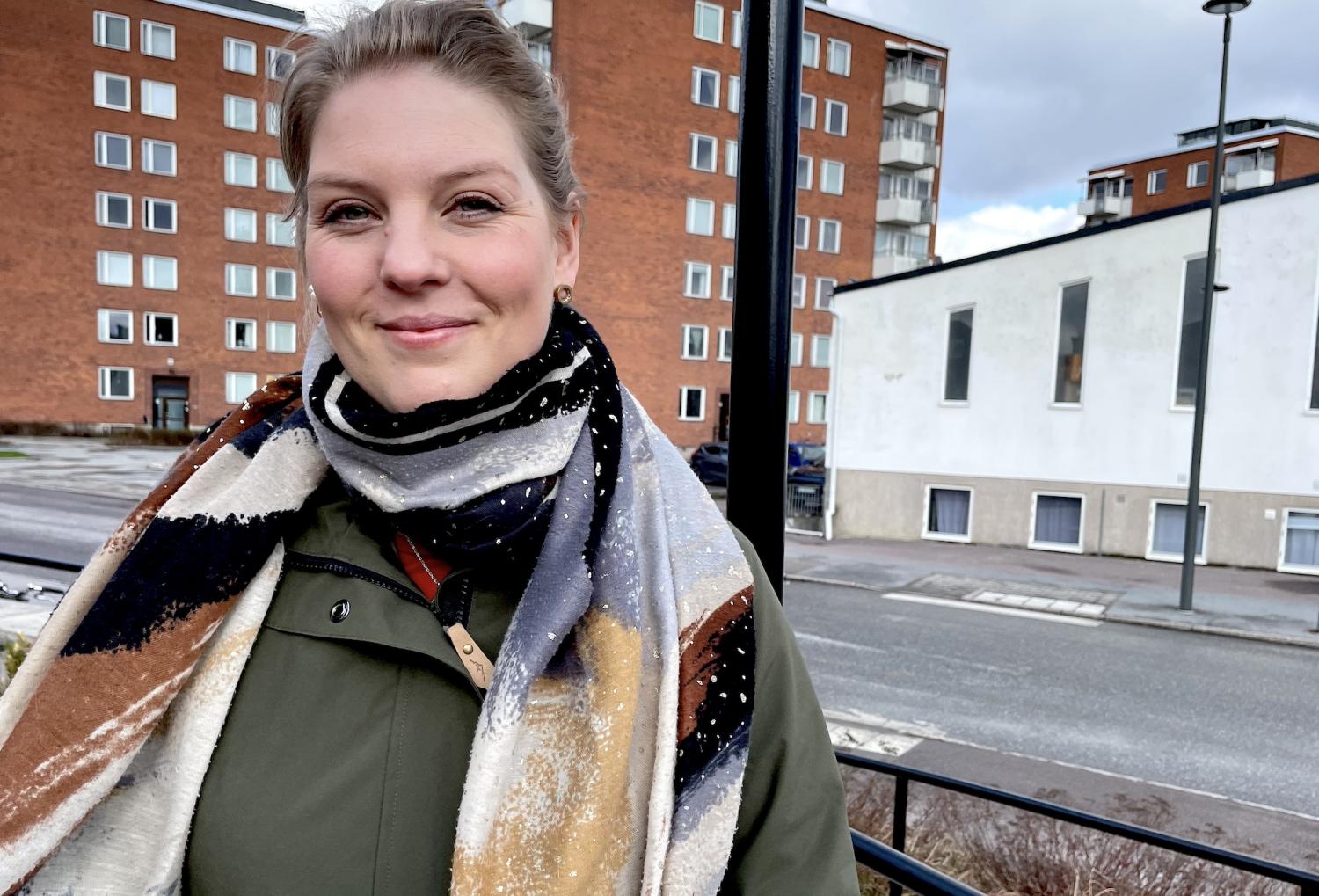 Kvinna i en färgglad halsduk och grön jacka som står på en balkong med flerbostadshus i bakgrunden en molnig dag.