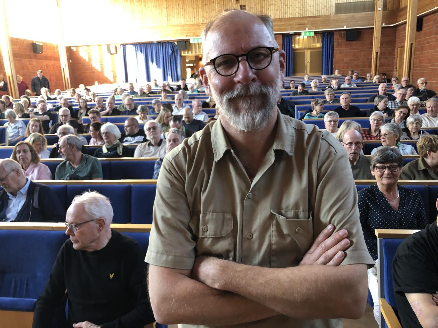 En man med glasögon och skägg står med armarna i kors framför en publik som sitter i ett auditorium av trä.