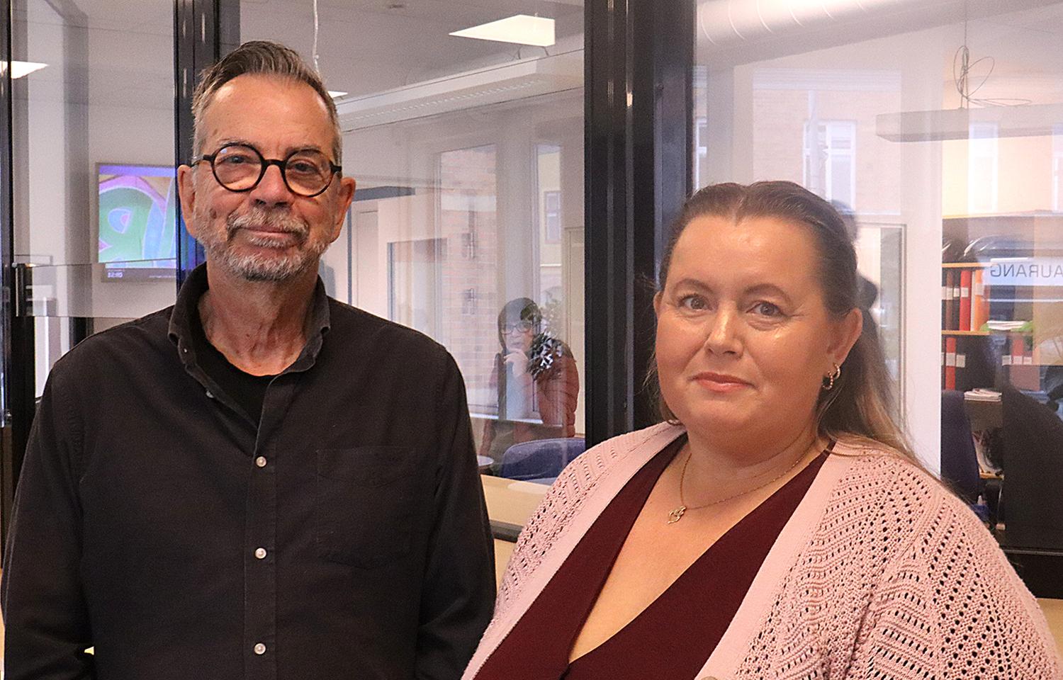 Två vuxna står inomhus i en kontorsmiljö och tittar in i kameran. Mannen bär glasögon och en mörk skjorta; kvinnan bär en rosa kofta och halsband.