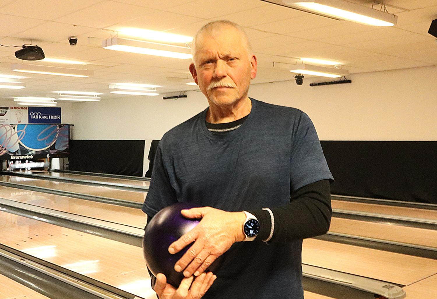 En äldre man står i en bowlinghall och håller i ett lila bowlingklot, klädd i en blå t-shirt, svarta byxor och en klocka. Bowlingbanor och käglor syns i bakgrunden.