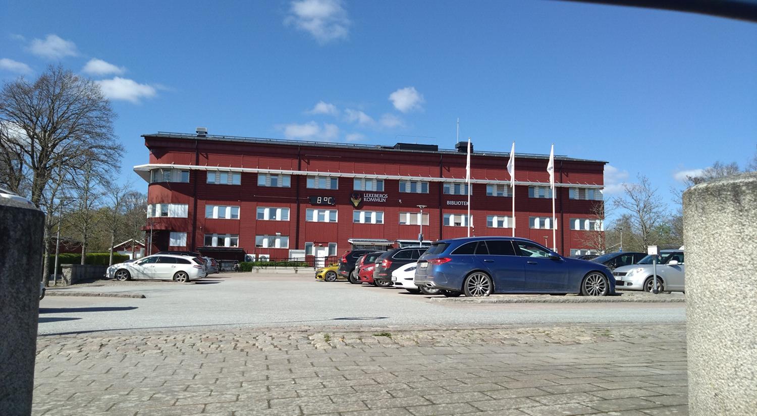 En röd trevåningsbyggnad med flera vita flaggor framför, en parkeringsplats full av bilar och en klarblå himmel i bakgrunden.