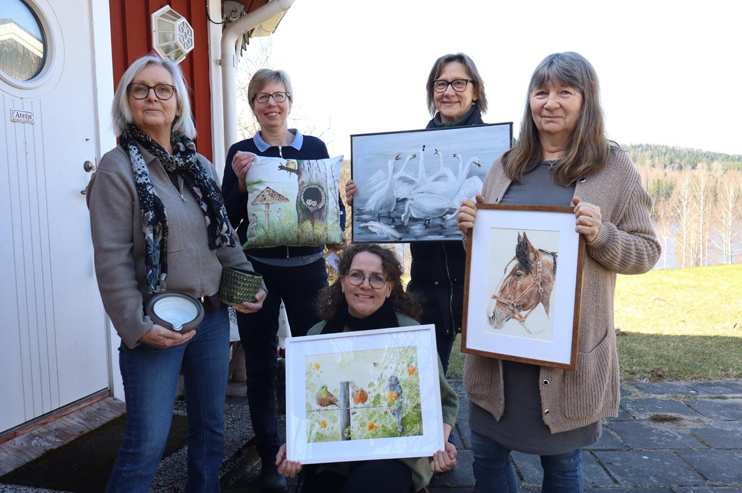 Fem kvinnor står utomhus vid ett hus, var och en med ett konstverk i handen, bland annat målningar av fåglar, en häst och en naturscen. I bakgrunden syns träd och ett vattendrag.