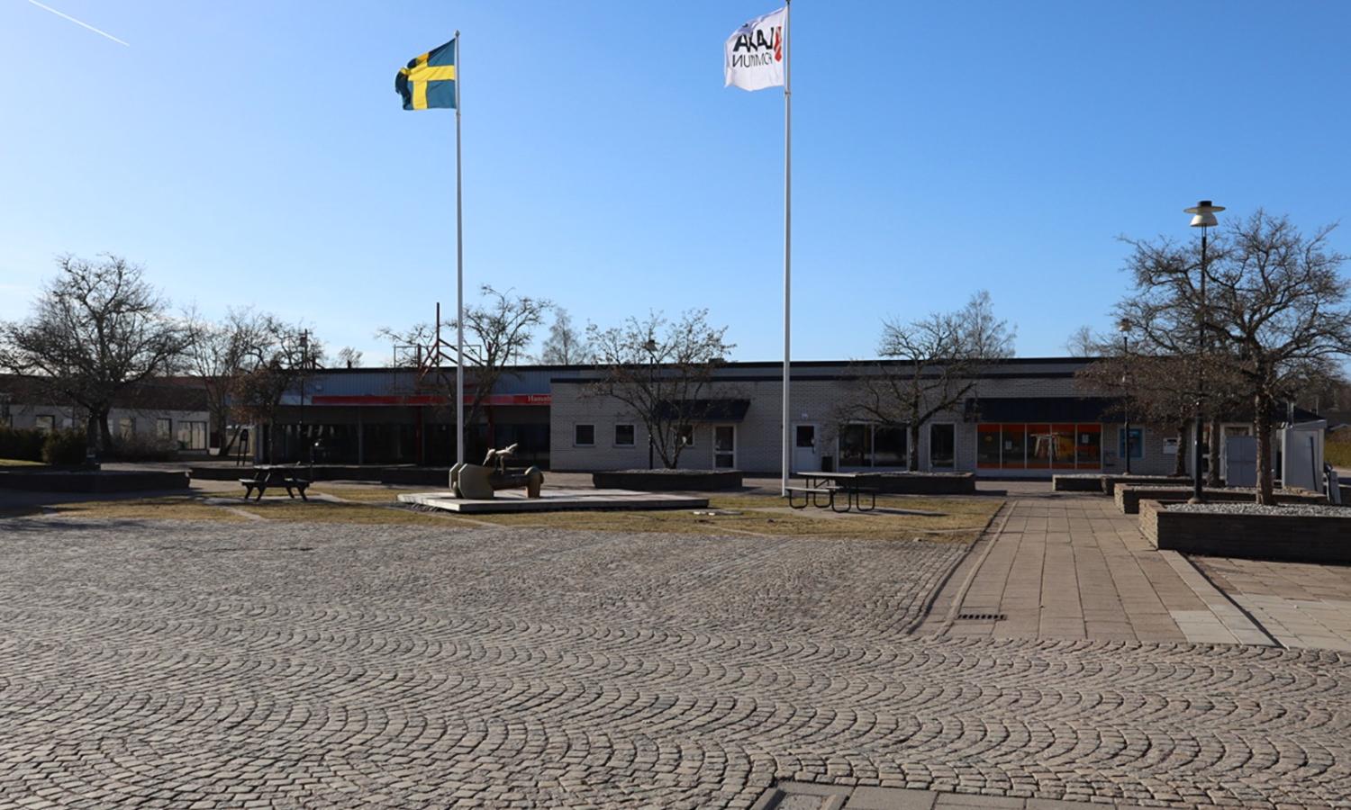 Ett asfalterat torg med två flaggstänger, en med en svensk flagga och en med en vit flagga, framför en låg byggnad under en klarblå himmel.