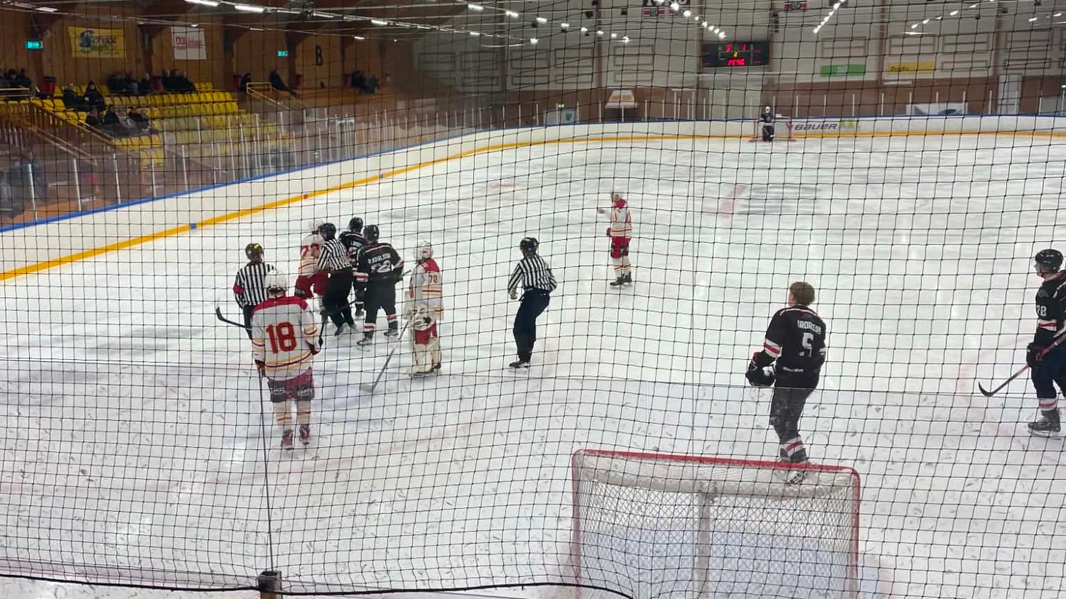 Ishockeyspelare samlas nära målet medan domarna ingriper, medan andra står och tittar på i närheten i en inomhushall, sedda genom skyddsnätet.