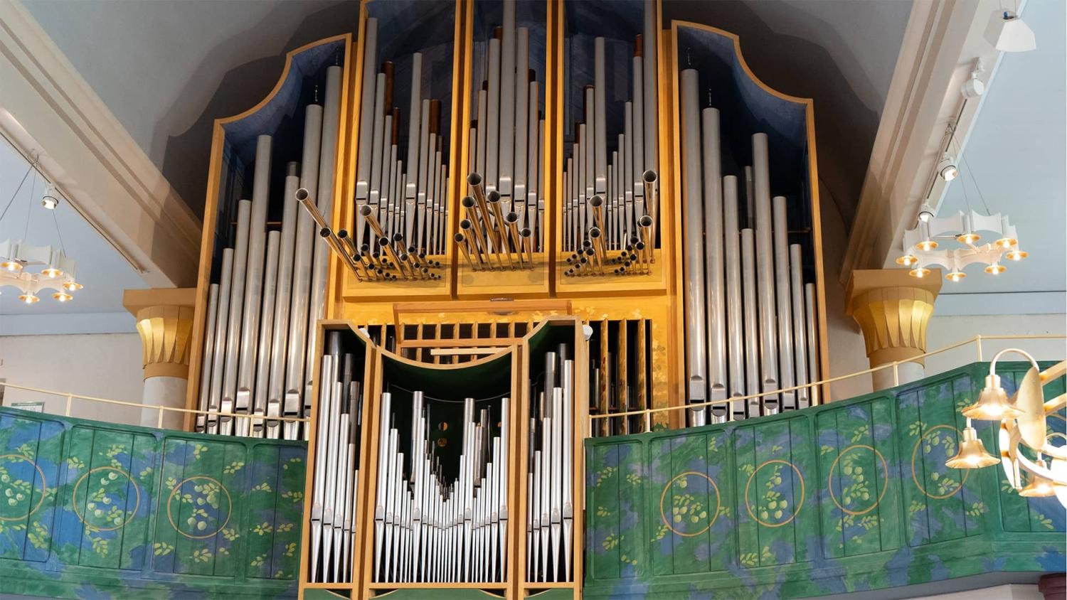 Stor piporgel med metallpipor och en dekorativ balkong i grönt och guld i en väl upplyst kyrkorum.