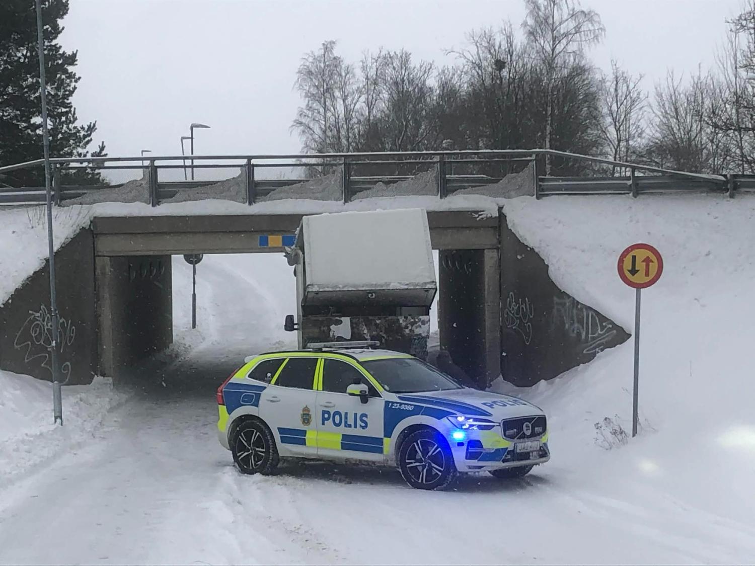 En polisbil blockerar en snöig väg där en lastbil har fastnat under en låg bro. Till höger syns en skylt om låg frigångshöjd.