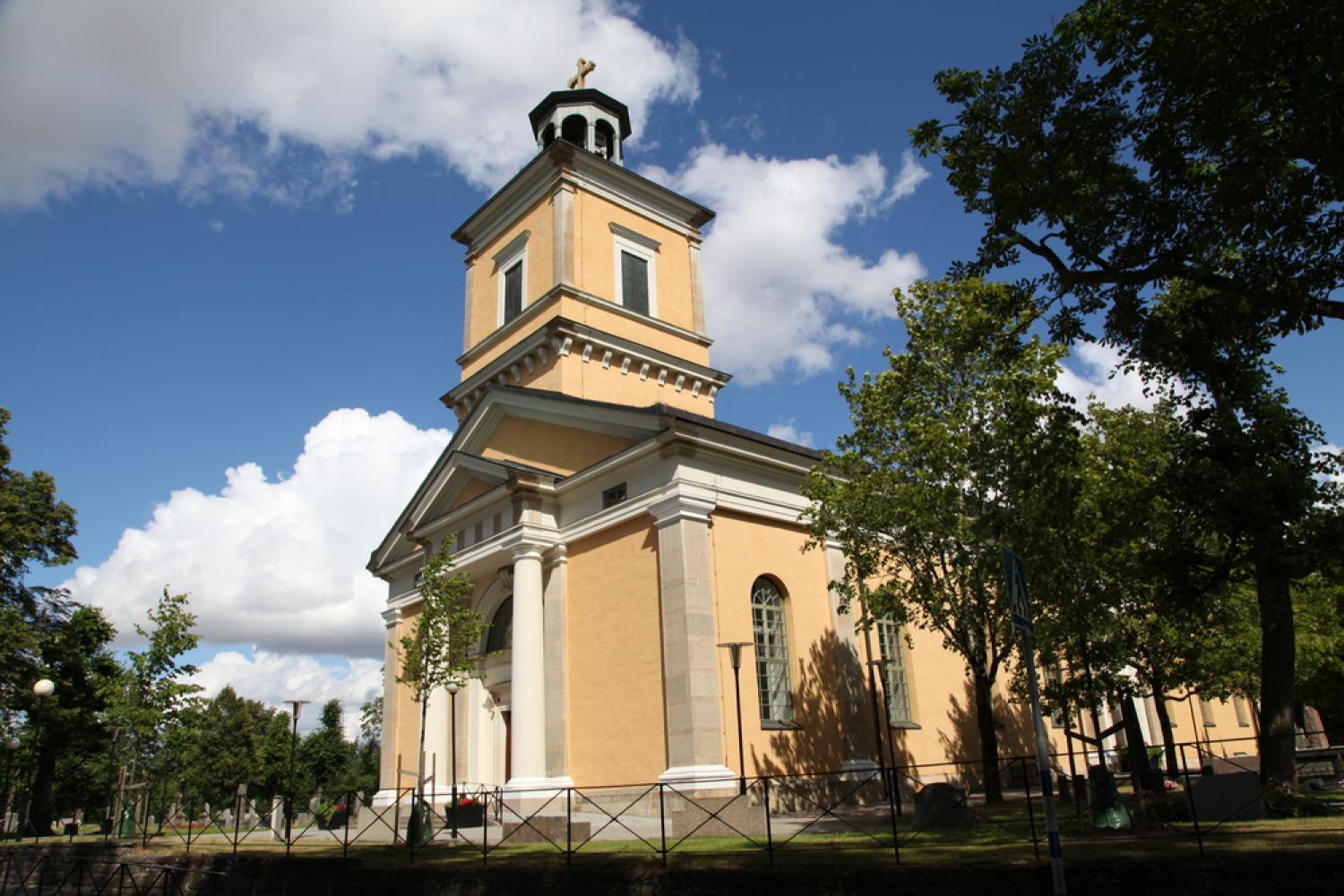 Gul kyrka med ett klocktorn med grönt tak, omgiven av träd och ett metallstaket, under en delvis molnig himmel.
