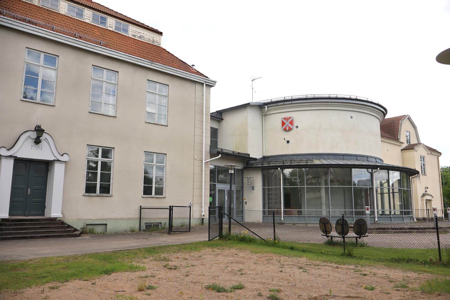 En beige tvåvåningsbyggnad med stora fönster och en modern glasutbyggnad, omgiven av ett stängsel av trådnät och en grusplan i förgrunden.