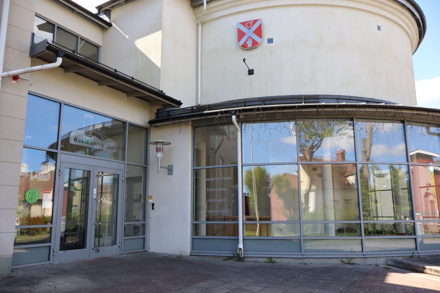 En modern byggnad med stora fönster, en glasentré och ett rödvitt vapen ovanför dörren; reflektioner av hus är synliga i glaset.