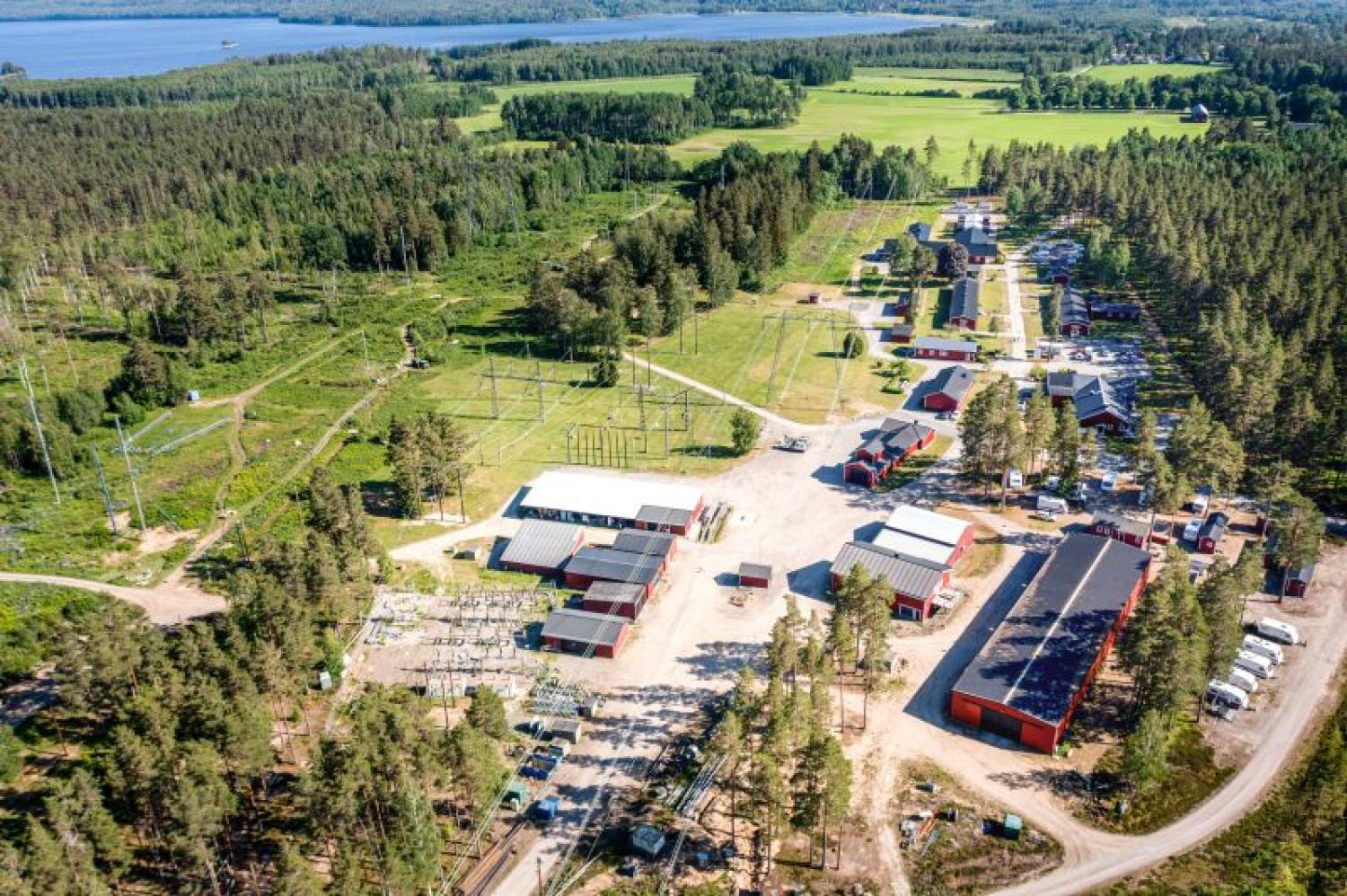 Flygfoto över en lantlig industrianläggning omgiven av skogar och fält, med flera byggnader och utrustning synlig, nära en vattenkropp i bakgrunden.