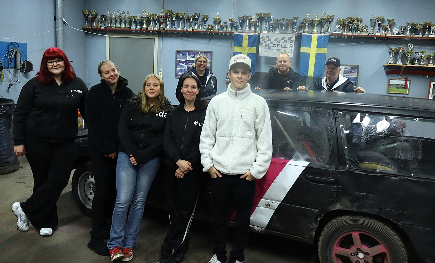 En grupp människor står bredvid en svart och rosa racerbil i ett garage, med pokaler på hyllor och svenska flaggor på väggen bakom dem.