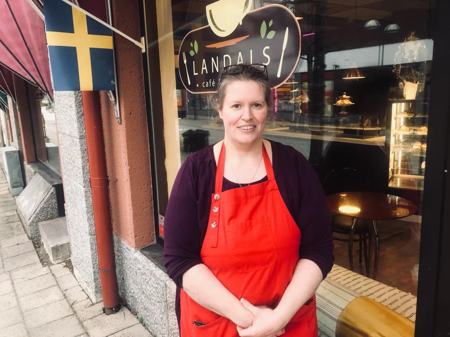 En person som bär ett rött förkläde står framför ett kafé med ett fönster bakom sig.