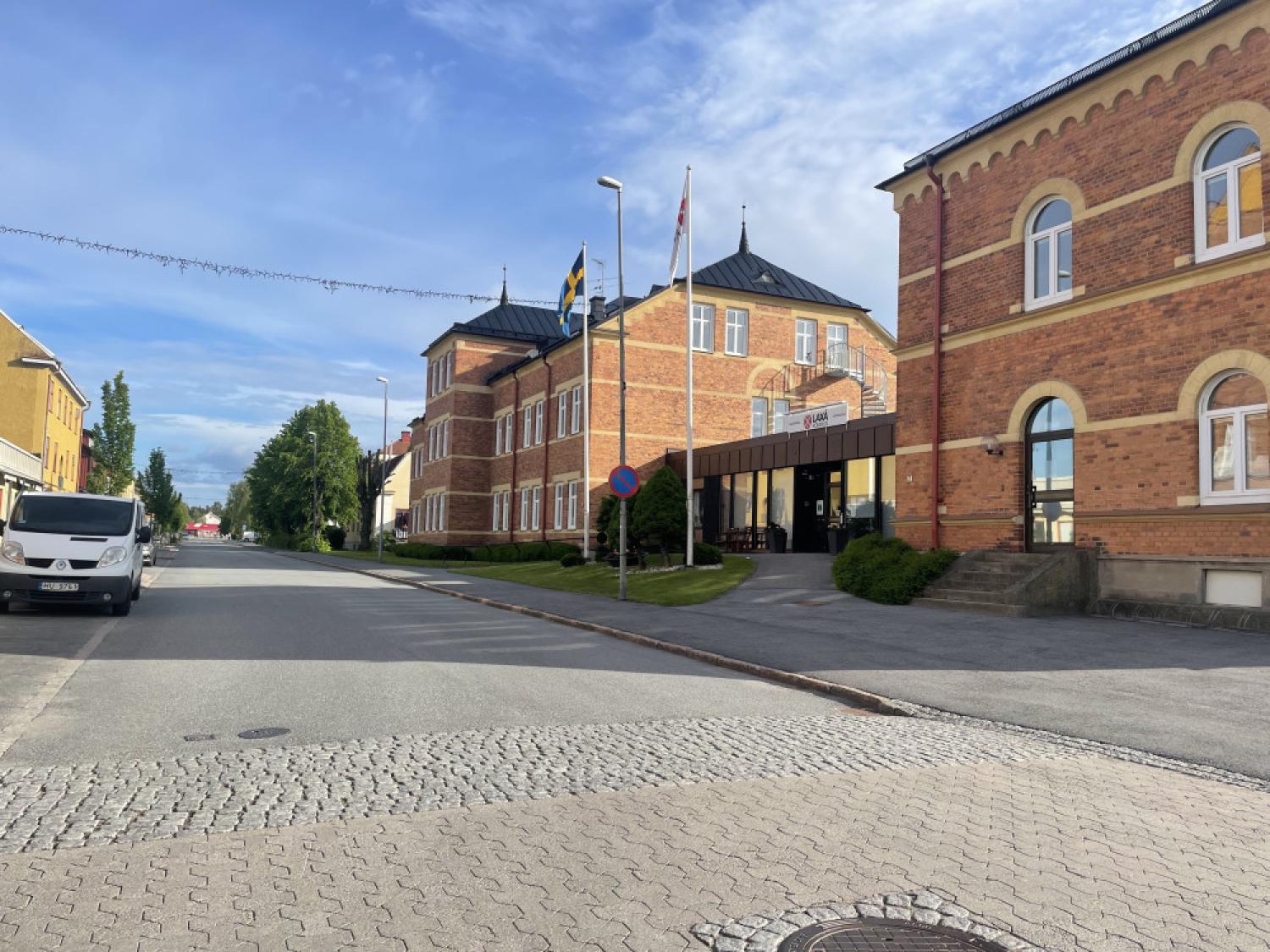 En byggnad i rött tegel med välvda fönster, omgiven av träd och flaggor. En skåpbil står parkerad på gatan och himlen är delvis molnig.