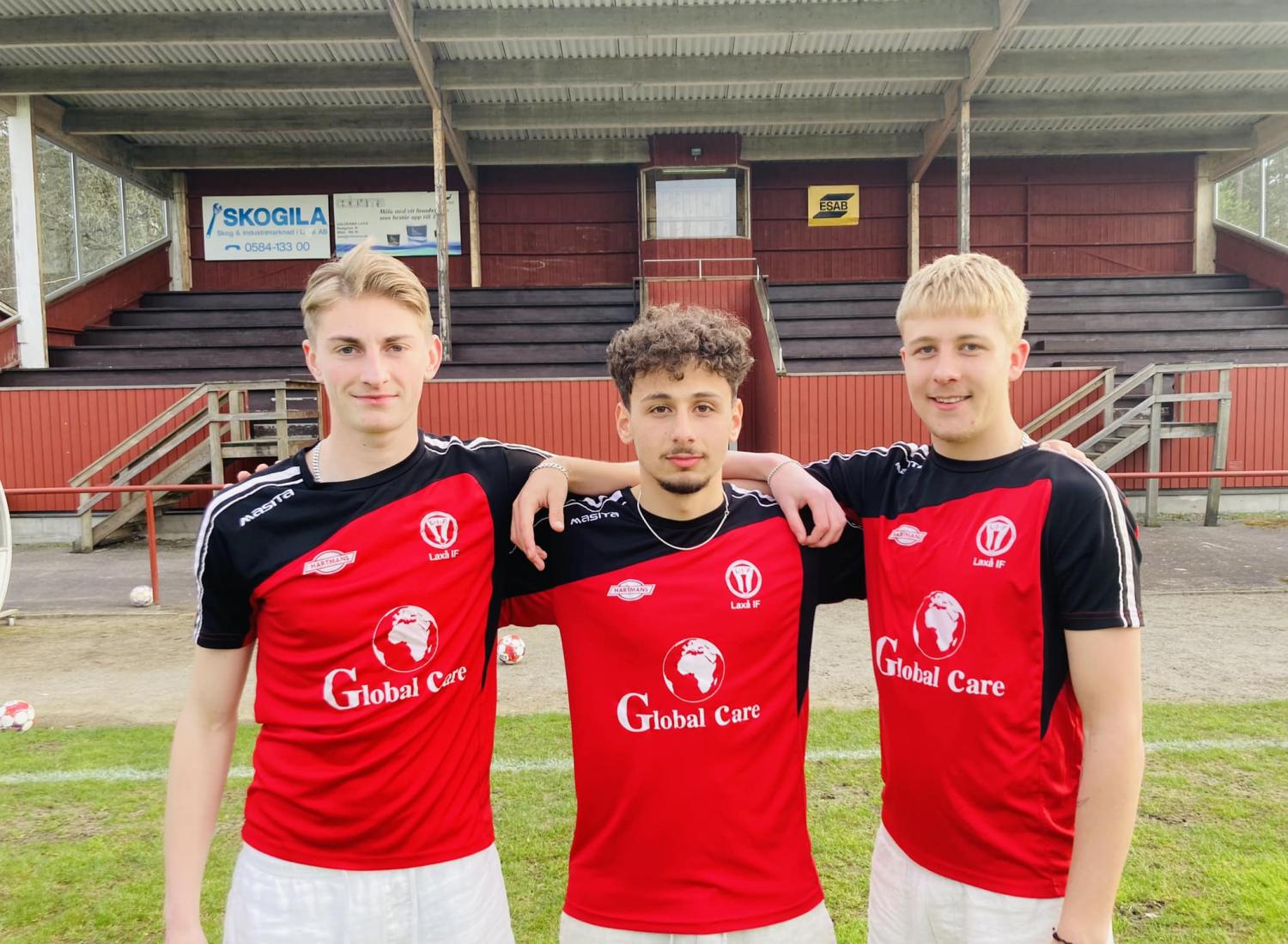 Tre unga män i röda och svarta fotbollströjor står på en plan med ett träställ i bakgrunden. Mannen i mitten har en arm runt var och en av de andra två männen.