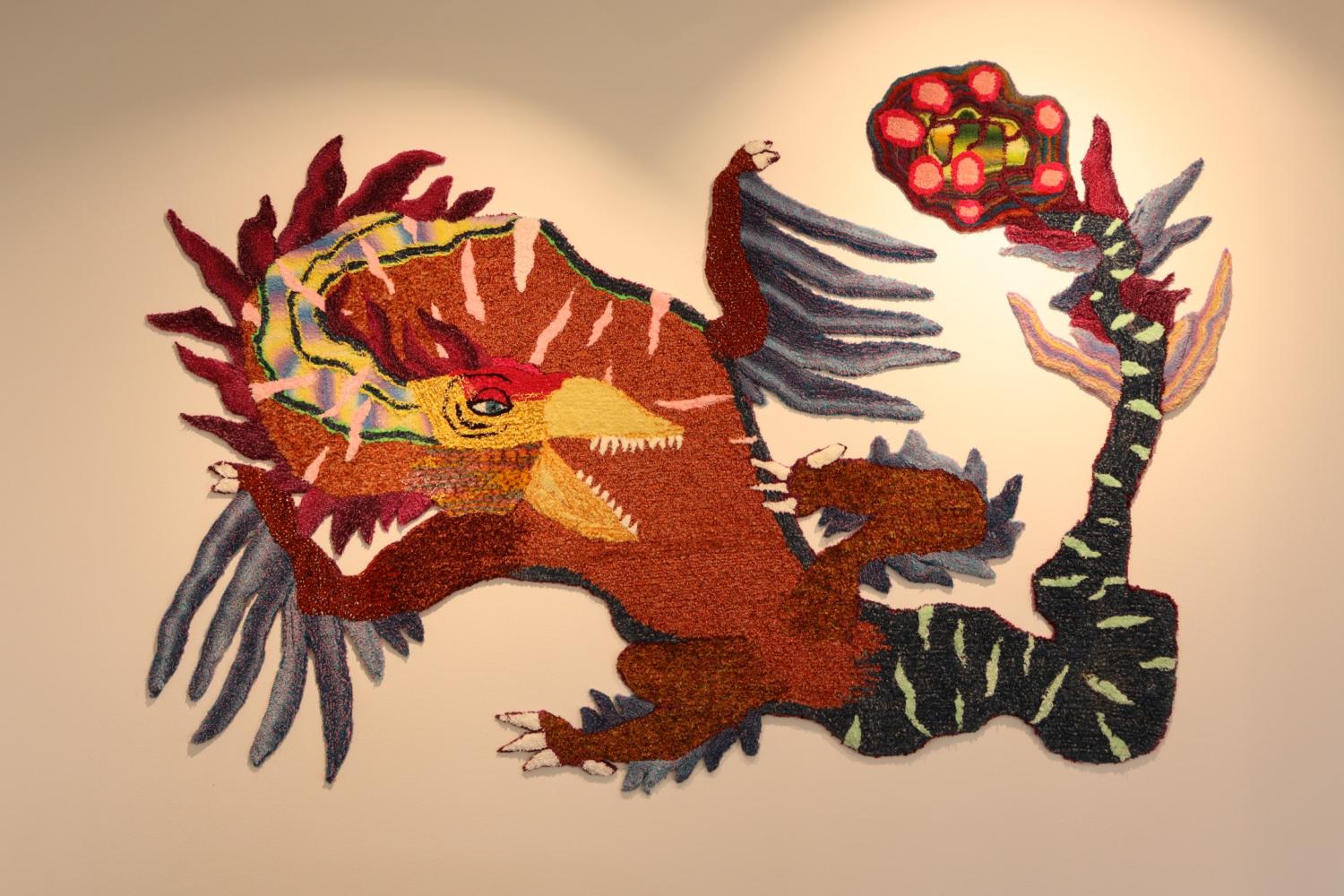 Textil väggkonst av en färgstark, abstrakt drakliknande varelse med överdrivna lemmar och en livlig svans, placerad på en ljus vägg med fokuserad belysning.