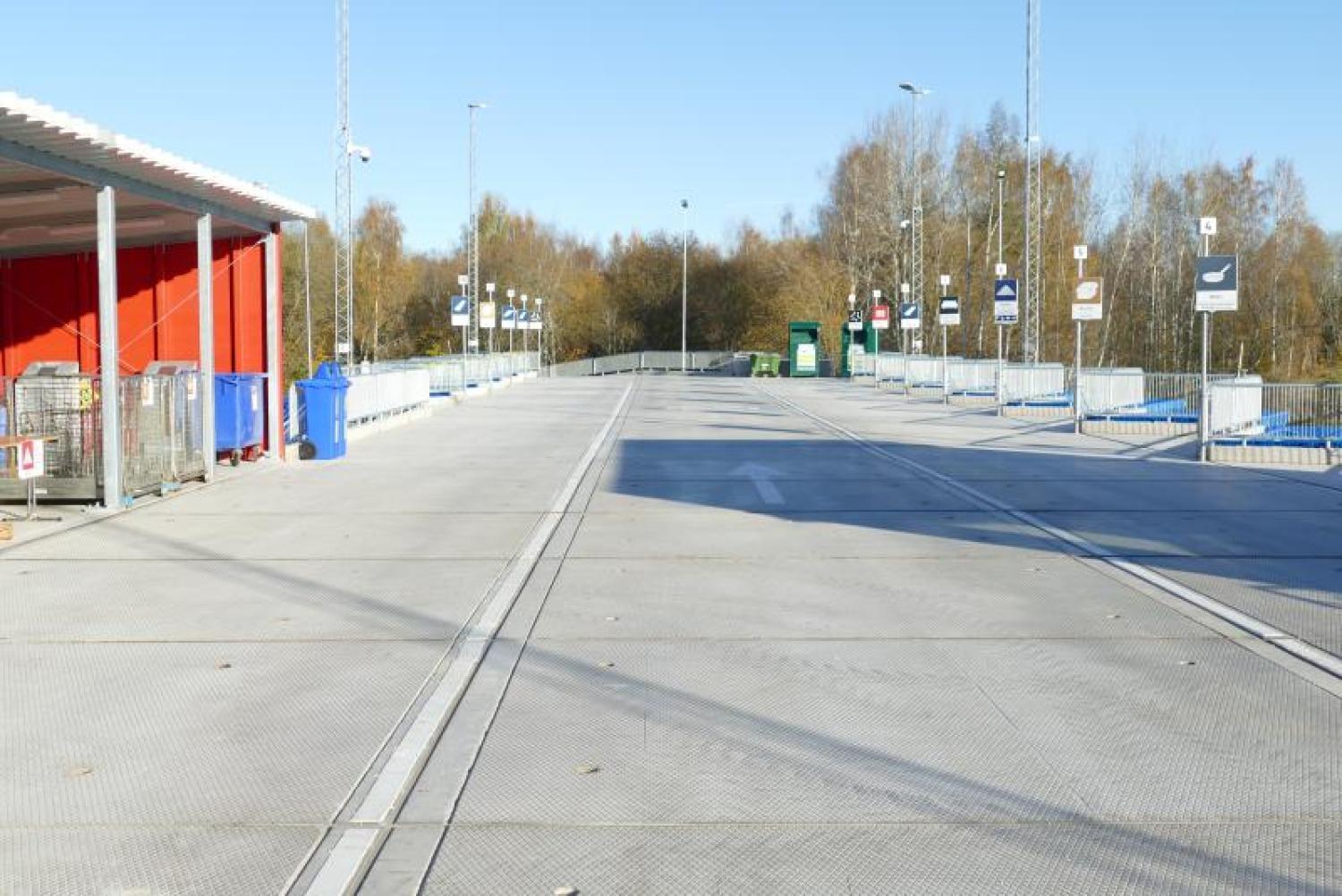 En ren, tom återvinningscentral utomhus med märkta soptunnor och skyltar, omgiven av träd och metallstaket under en klar himmel.