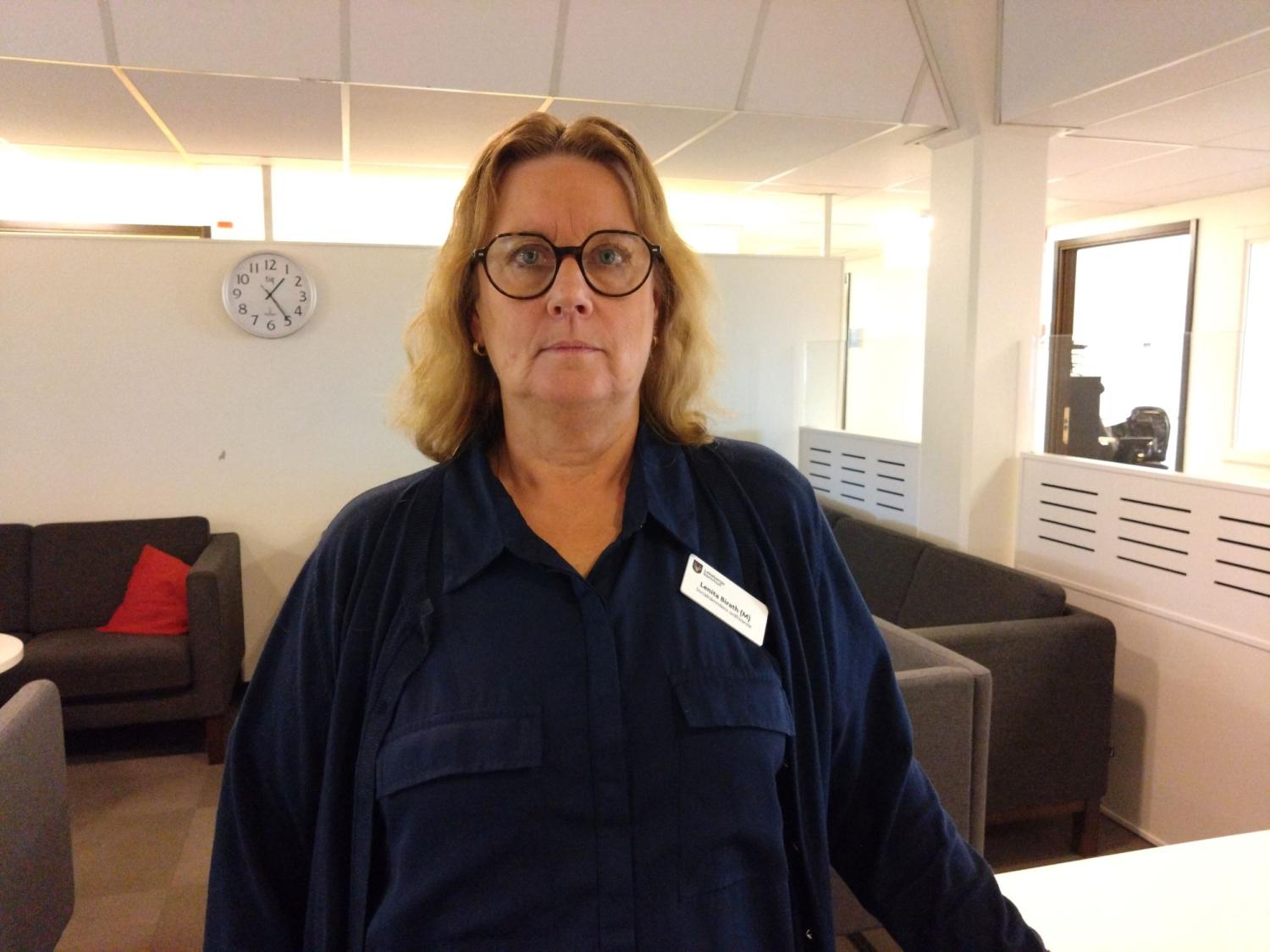 En person med glasögon står på ett kontor och bär en namnskylt. En klocka på väggen visar att klockan är något efter 8:00. Det finns stolar och ett bord i bakgrunden.