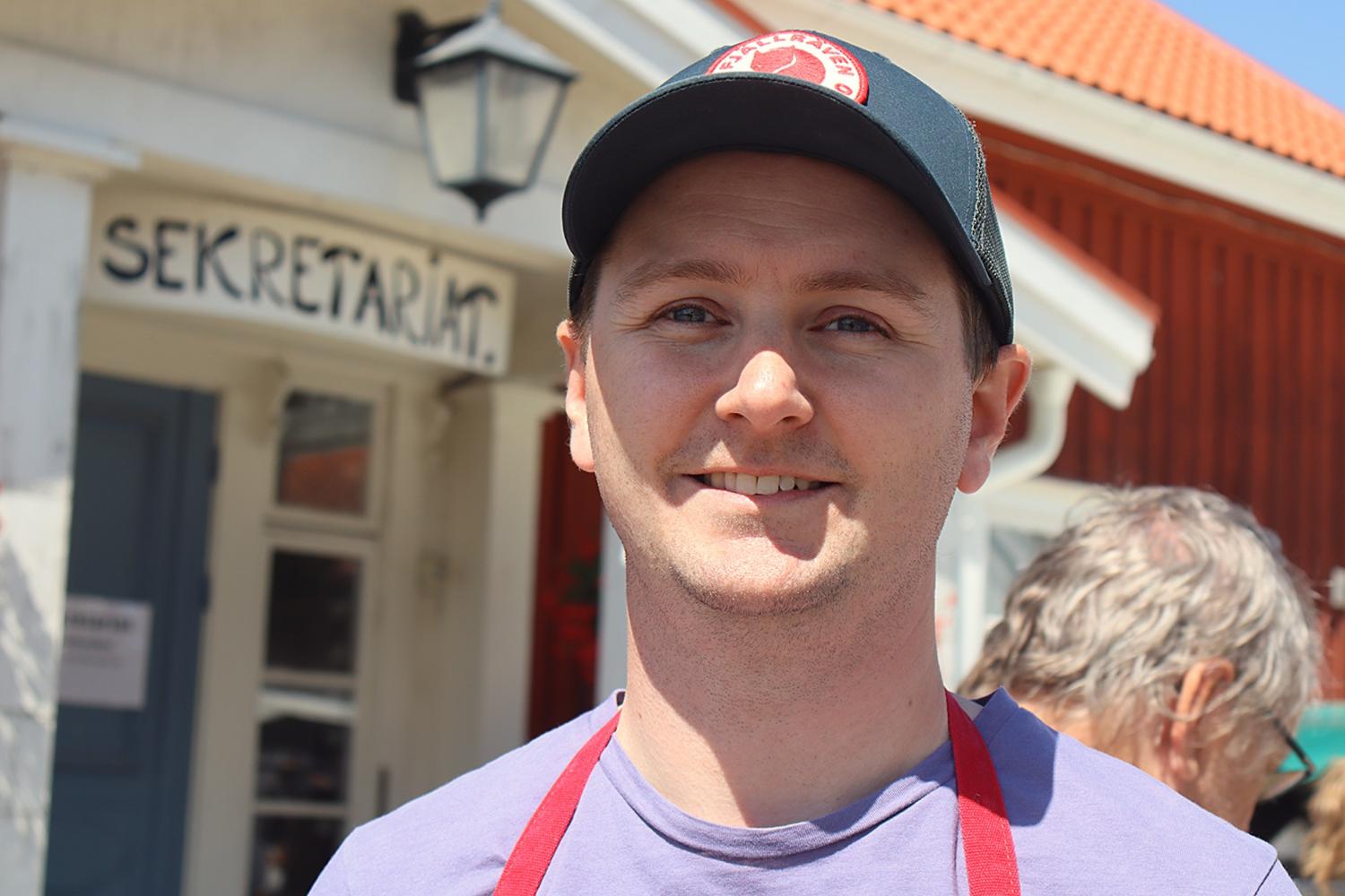 En man med svart keps och rött förkläde står ute i solskenet och ler mot kameran. I bakgrunden syns en byggnad med en skylt där det står ”SEKRETARIAT”.