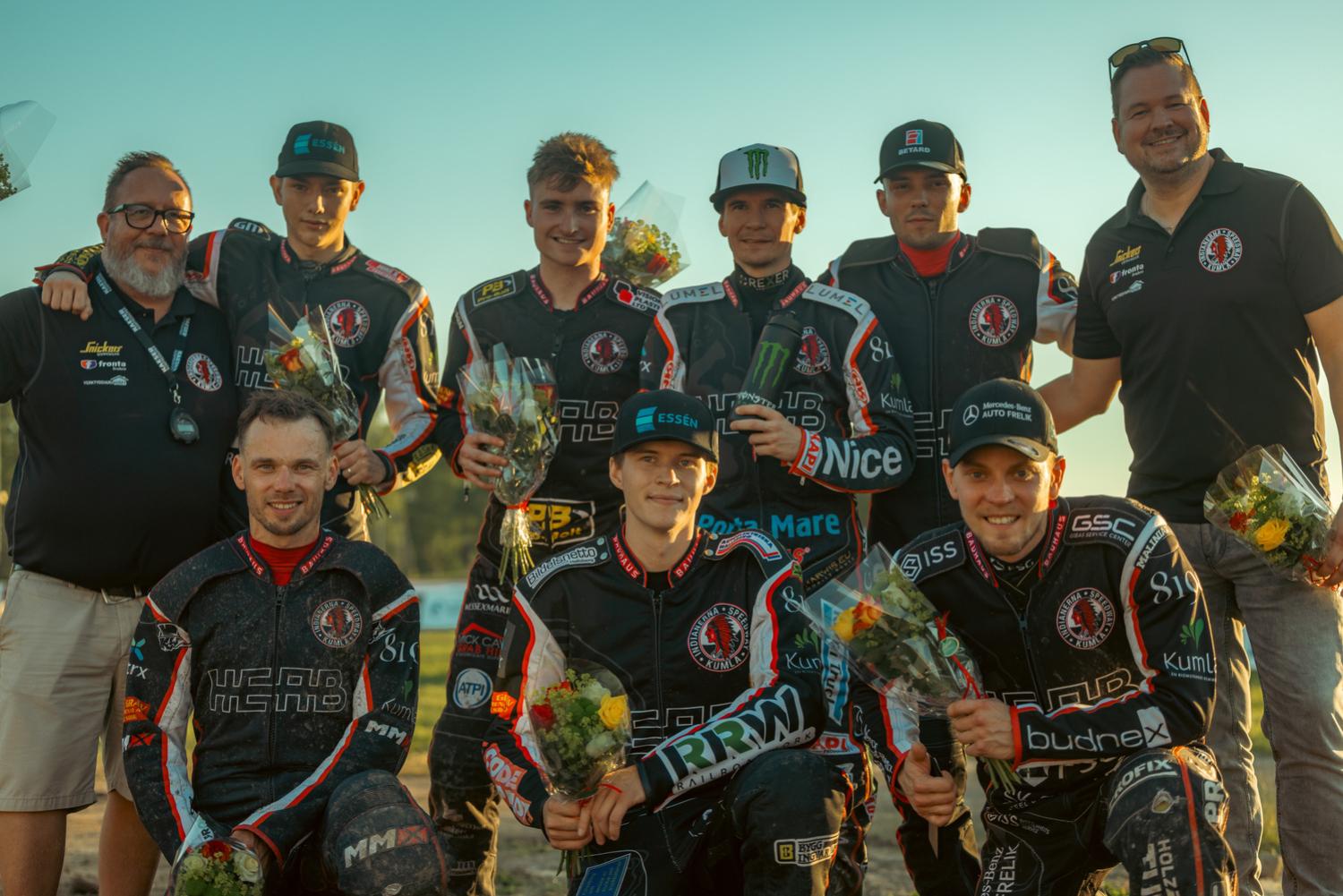 En grupp på åtta personer, inklusive motocrossteammedlemmar i racingutrustning som håller i blombuketter och två män i vardagskläder, poserar för ett foto utomhus under solnedgången.